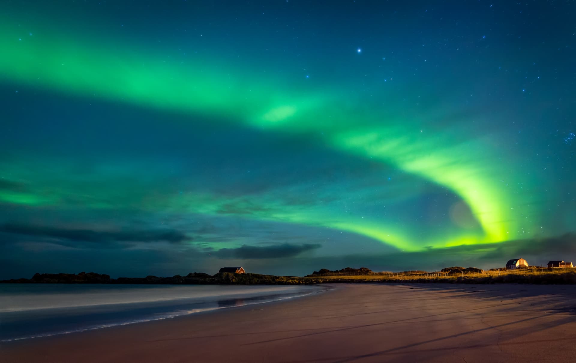 Aurora boreal verde iluminando o céu estrelado sobre uma praia e casas iluminadas